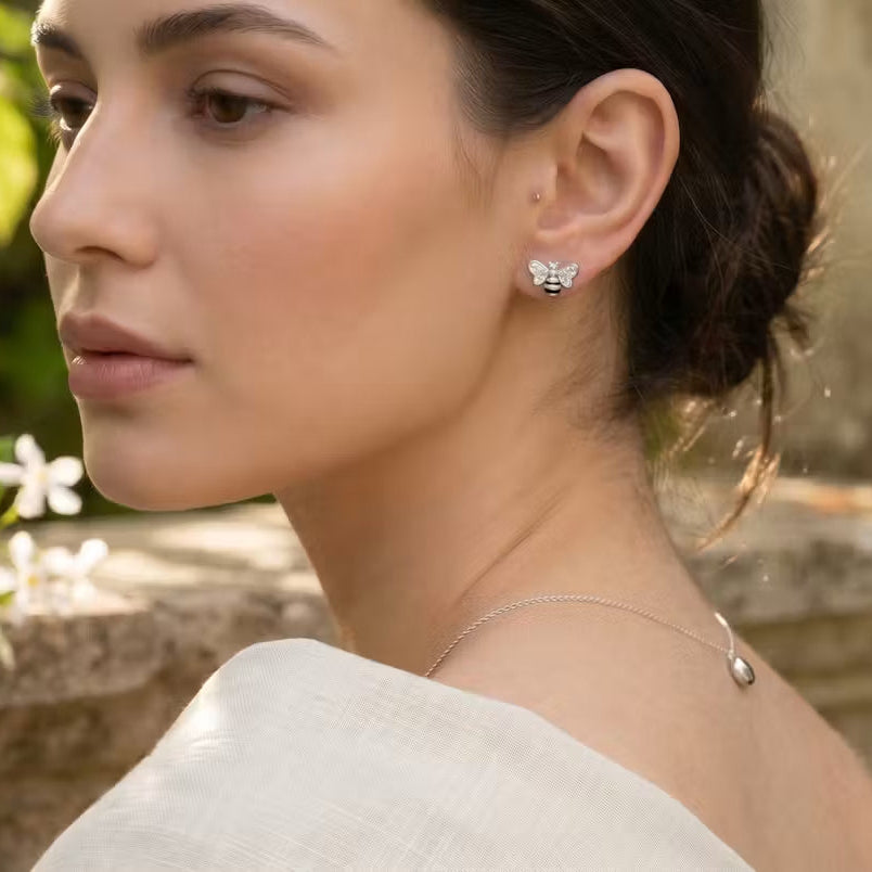A woman in profile wearing a delicate silver necklace and a small silver butterfly stud earring, set against a soft-focus outdoor background of green foliage and a stone wall.